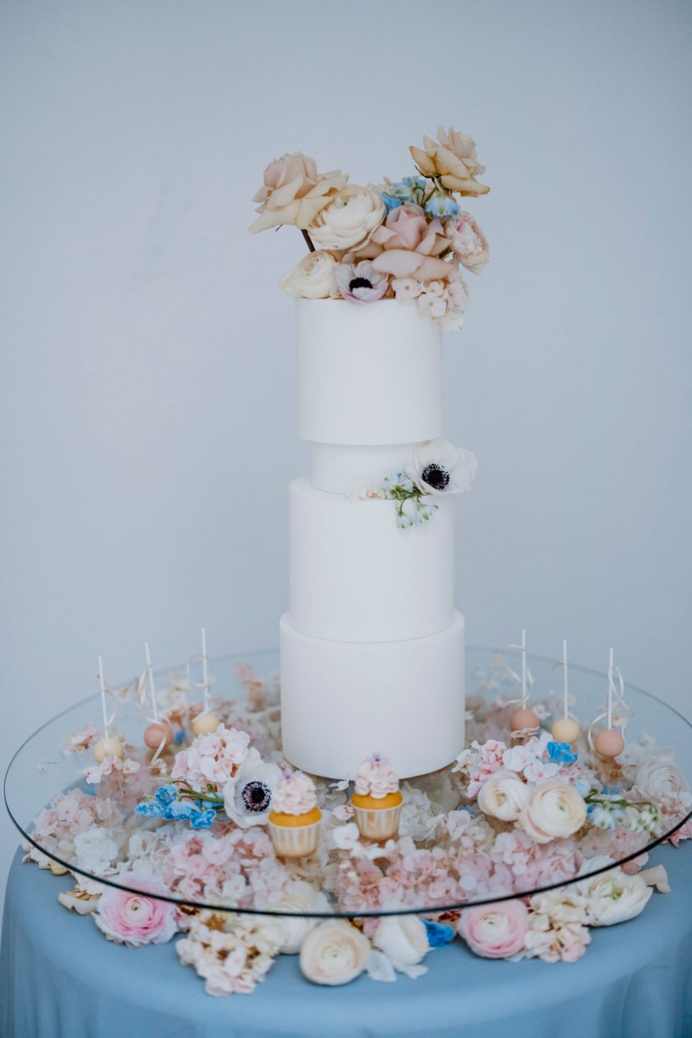 Hochzeitswahn - Eine elegante dreistöckige weiße Hochzeitstorte, geschmückt mit pastellfarbenen Blumen, umgeben von einer Reihe wunderschön dekorierter Cupcakes und floraler Akzente, präsentiert auf einem Glastisch.