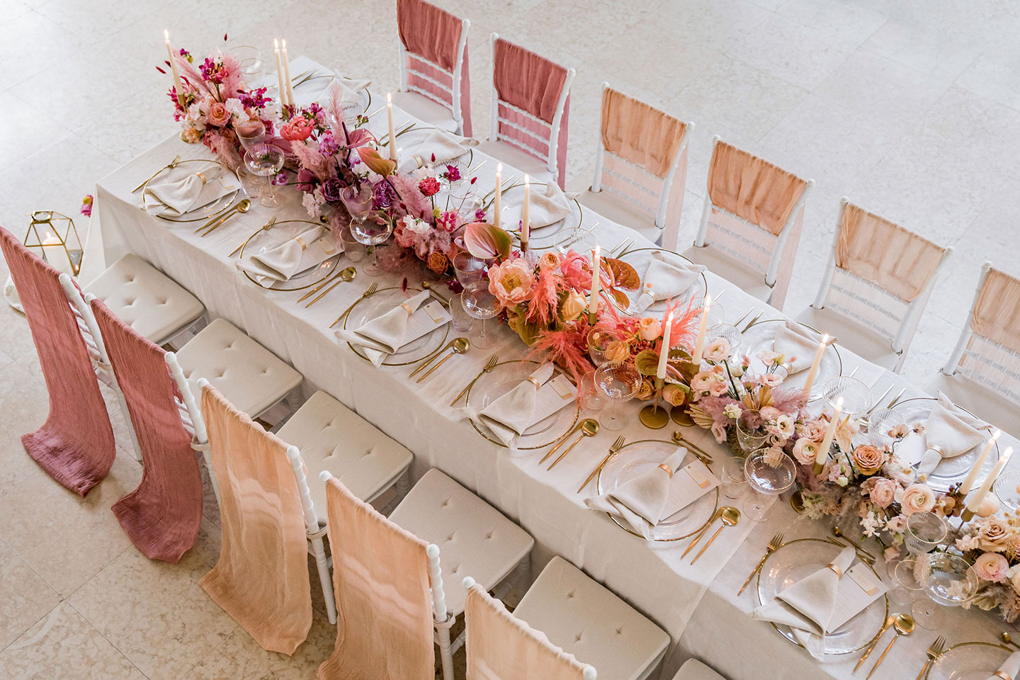 Hochzeitswahn - Ein elegant gedeckter Hochzeitsempfangstisch mit Blumenarrangements in Rosa- und Pfirsichtönen, goldenem Besteck und feinem Glasgeschirr, ergänzt durch anmutige Chiffon-Stuhlvorhänge in einem anspruchsvollen Veranstaltungsort.