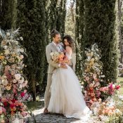 Hochzeitswahn - Ein Paar umarmt sich an seinem Hochzeitstag, umgeben von üppigem Grün und farbenfrohen Blumenarrangements, und genießt die ruhige, sonnenbeschienene Gartenkulisse.