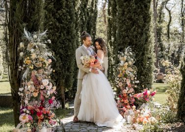 Hochzeitswahn - Ein Paar umarmt sich an seinem Hochzeitstag, umgeben von üppigem Grün und farbenfrohen Blumenarrangements, und genießt die ruhige, sonnenbeschienene Gartenkulisse.