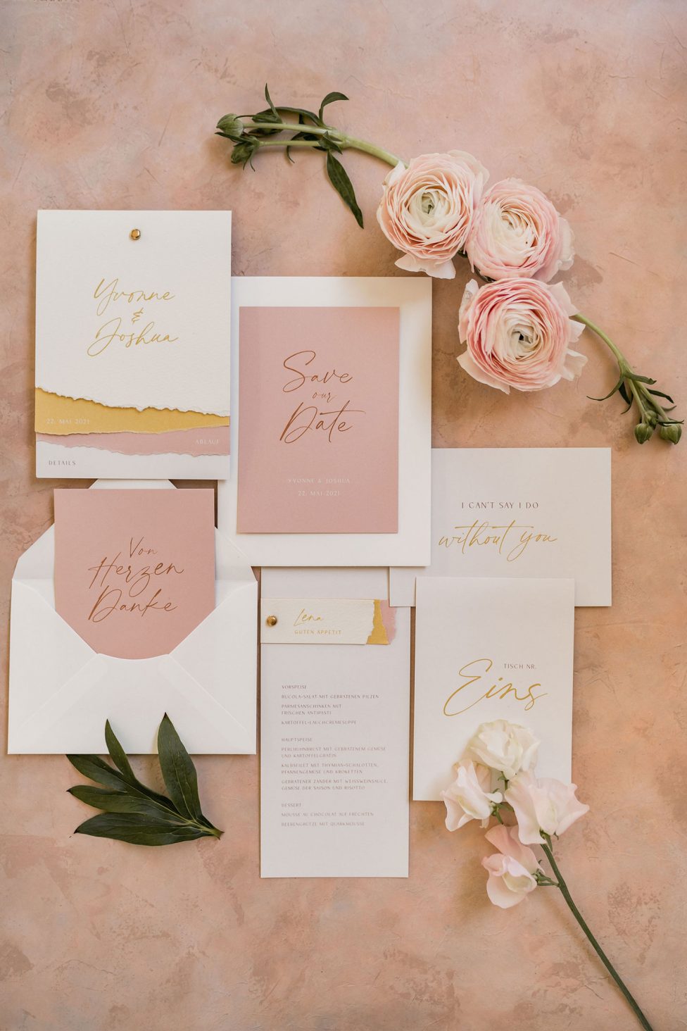 Hochzeitswahn - Ein elegantes Hochzeitspapeterie-Set, einschließlich Einladungen und Save-the-Date-Karten, geschmückt mit goldenen Akzenten, auf einem rosigen Hintergrund angeordnet, ergänzt durch zarte Ranunkelnblüten und Grünpflanzen.