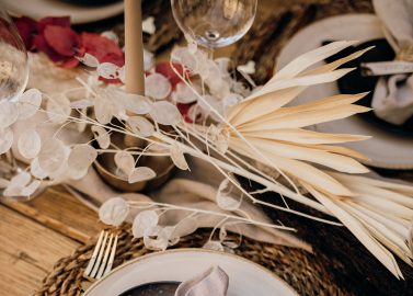 Hochzeitswahn - Ein elegant gedeckter Tisch mit einem rustikalen geflochtenen Unterteller, einem dunkel mattierten Teller, goldenem Besteck, einer Leinenserviette und einem personalisierten, mit Bindfaden befestigten Namensschild für ein anspruchsvolles Dinner-Event.