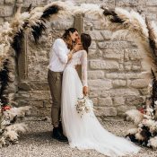 Hochzeitswahn - Ein Paar küsst sich an seinem Hochzeitstag zärtlich, eingerahmt von einem Bohème-Bogen aus Pampasgras und Blumen. Die Braut trägt ein Spitzenkleid und der Bräutigam Grau als Zeichen ihrer liebevollen Verbindung.