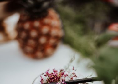 Hochzeitswahn - Eine Nahaufnahme eines Glases mit einem geschichteten, cremigen, weißen Getränk, garniert mit rosa Streuseln, einem Kräuterzweig und einem schwarzen Strohhalm, mit einer verschwommenen Ananas im Hintergrund.