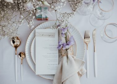 Hochzeitswahn - Elegantes Tischgedeck mit einem weißen Teller mit Goldrand, silbernem Besteck und einer mit Schnur zusammengebundenen und mit violetten Blumen geschmückten Leinenserviette. Eine gedruckte Speisekarte, feines Glasgeschirr und sanfte Blumendekorationen ergänzen das minimalistische Thema.