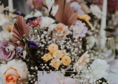 Hochzeitswahn - Eine elegante Tischdekoration für eine Hochzeit, deren Tafelaufsatz aus üppigen, bunten Blumen und zartem Schleierkraut besteht. Im Vordergrund ist ein transparentes „Table One“-Schild zu sehen, flankiert von einer Kerze und einer kleinen bunten Geschenkbox.