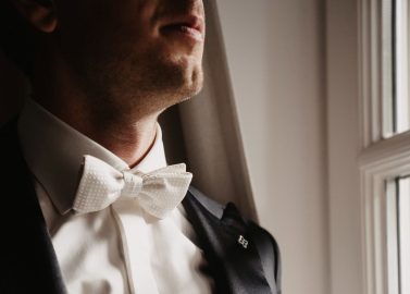 Hochzeitswahn - Eine Person steht neben einem Fenster im Schloss Gartrop. Sie trägt einen dunklen Anzug, ein weißes Oberhemd und eine weiße Fliege. Das Licht, das durch das Fenster fällt, hebt die Kleidung der Person und den unteren Teil ihres Gesichts hervor und verleiht ihr einen Hauch von Eleganz, der perfekt zu einer Schlosshochzeit im Ruhrgebiet passt.