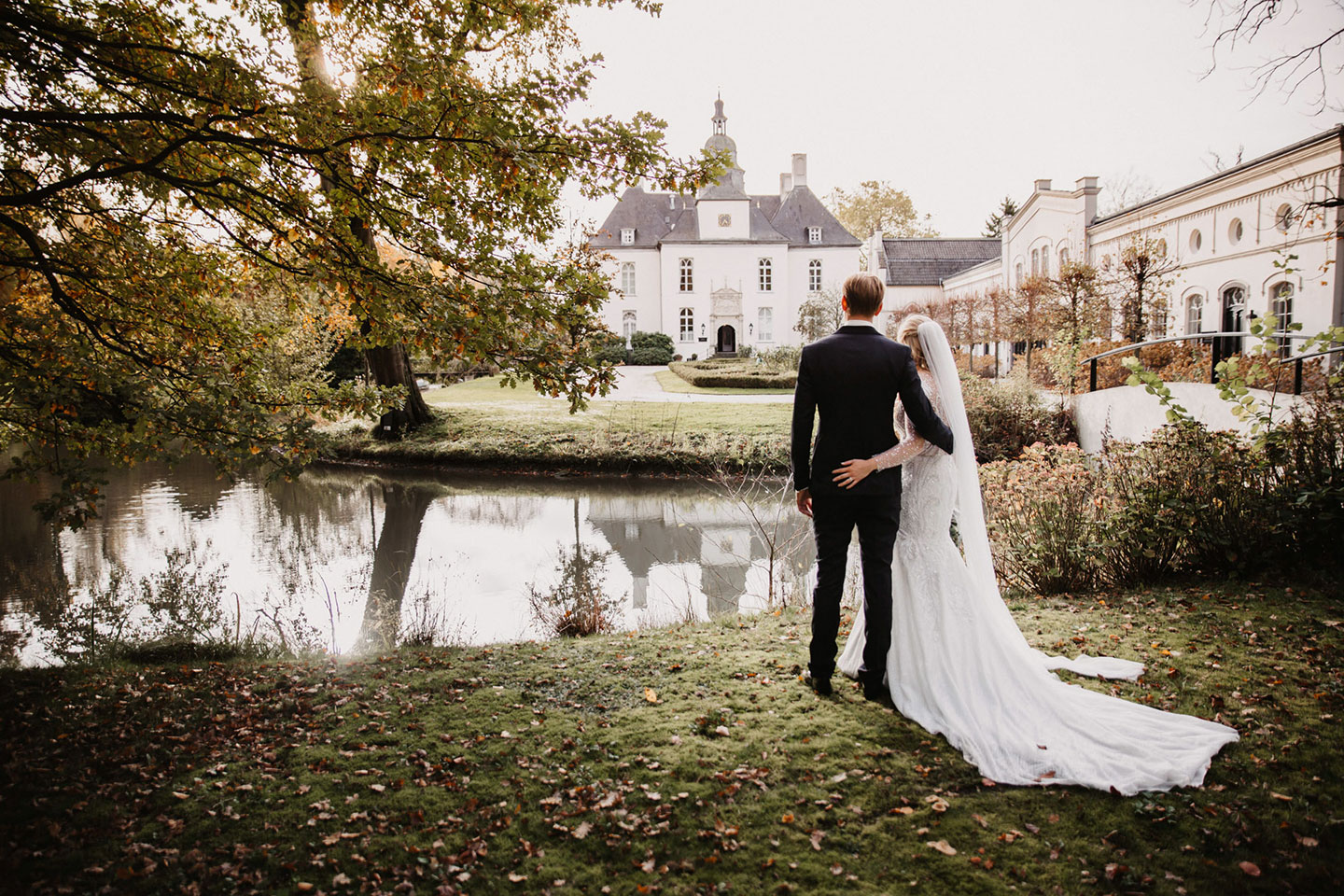 Hochzeitswahn - Braut und Bräutigam stehen an einem Teich vor Schloss Gartrop, einem eleganten Gebäude im Ruhrgebiet. Die Braut, in einem weißen Kleid mit langer Schleppe und Schleier, und der Bräutigam, in einem dunklen Anzug, sind von hinten zu sehen, wie sie einander umarmen. Bäume und Laub umgeben den Schauplatz ihrer Schlosshochzeit.