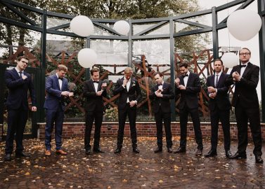 Hochzeitswahn - Eine Gruppe von acht Männern in Anzügen steht draußen unter großen weißen Luftballons vor Schloss Gartrop. Sie richten ihre Uhren und Manschetten, während sie sich im Halbkreis gegenüberstehen. Der Boden ist mit gelben Blättern bedeckt, im Hintergrund sind Bäume und ein Metallrahmen zu sehen, was eine malerische Schlosshochzeitsszene im Ruhrgebiet ergibt.