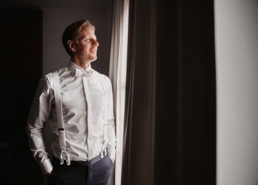 Hochzeitswahn - Ein Mann in weißem Hemd und weißen Hosenträgern steht neben einem Fenster im Schloss Gartrop, durch das Licht hereinströmt. Er hat die Hände in den Taschen und blickt mit einem leichten Lächeln hinaus. Der Raum, wahrscheinlich Teil der reizvollen Kulisse für eine Schlosshochzeit im Ruhrgebiet, scheint schwach beleuchtet.