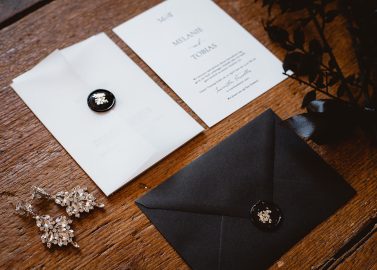 Hochzeitswahn - Elegantes Hochzeitseinladungsset auf einer Holzoberfläche. Enthält eine weiße Einladungskarte mit Namen, einen schwarzen Umschlag mit Siegel und daneben eine florale Haarnadel. Innviertler-Versailles