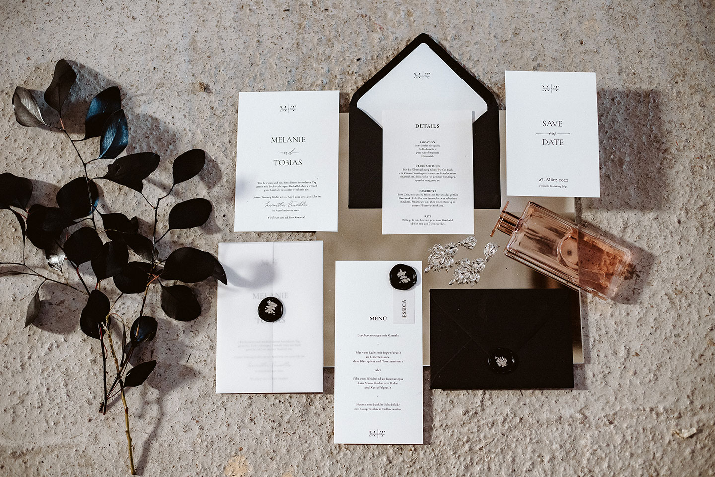 Hochzeitswahn - Eine Auswahl eleganter Hochzeitspapeterie, einschließlich Einladungen und Menüs, präsentiert mit einem schwarzen Umschlag, Wachssiegel und natürlichen Dekorationen auf einer strukturierten Betonoberfläche. Innviertler-Versailles
