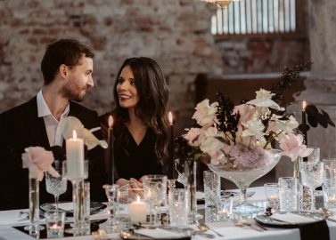Hochzeitswahn - Ein Paar lächelt und unterhält sich bei einem romantischen Abendessen mit Kerzen und einem großen Blumengesteck in einem schwach beleuchteten rustikalen Raum. Innviertler-Versailles