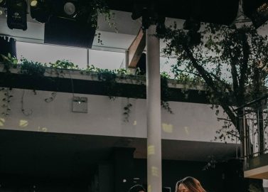 Hochzeitswahn - Ein fröhliches Paar tanzt bei einer lebhaften Indoor-Veranstaltung, wobei der Mann die Frau hochhebt, die breit lächelt. Im Hintergrund spielt eine Band vor einer Kulisse aus Lichterketten und Grünpflanzen. Villa-Flora