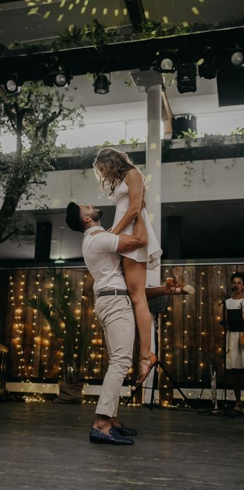 Hochzeitswahn - Ein Mann hebt eine Frau während eines Tanzes bei einer Hochzeitsfeier hoch, umgeben von Lichterketten und Gästen. Die Szene spielt in einem Innenbereich, im Hintergrund ist eine Live-Band zu sehen. Villa-Flora