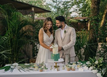 Hochzeitswahn - Ein Paar lächelt bei seiner Hochzeitsfeier, während es eine mehrstöckige Torte anschneidet, umgeben von einer tropischen Dekoration mit üppigem Grün und Lichterketten. Villa-Flora