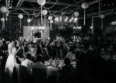 Hochzeitswahn - Ein Paar umarmt sich zärtlich auf der Tanzfläche inmitten eines rustikalen Scheunenempfangs, der von hängenden Laternen beleuchtet wird. Die Gäste sitzen an mit Blumendekorationen geschmückten Tischen und genießen die festliche Atmosphäre.