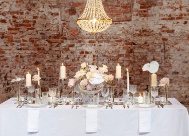 Hochzeitswahn - Elegante Esszimmereinrichtung mit weißer Tischdecke, Kristallgläsern und goldenem Besteck. Ein Tafelaufsatz mit blassrosa Blumen und weißen Kerzen, alles unter einem Kristallleuchter vor einer rustikalen Backsteinwand. Innviertler-Versailles