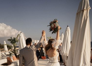 Hochzeitswahn - Ein frisch verheiratetes Paar verlässt freudig seine Hochzeitszeremonie unter strahlendem Himmel, während die Gäste fröhlich ihre Hände heben und so einen Torbogen schaffen, um die Hochzeit zu feiern.
