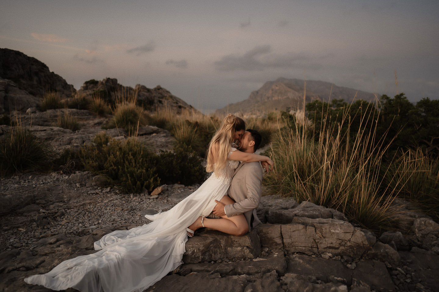 Hochzeitswahn - Ein Paar teilt einen zärtlichen Moment auf einem felsigen Gelände in der Dämmerung, wobei die Frau in einem fließenden Kleid auf dem Schoß des Mannes sitzt und sich umarmt, vor der Kulisse entfernter Berge unter einem sanften, düsteren Himmel.