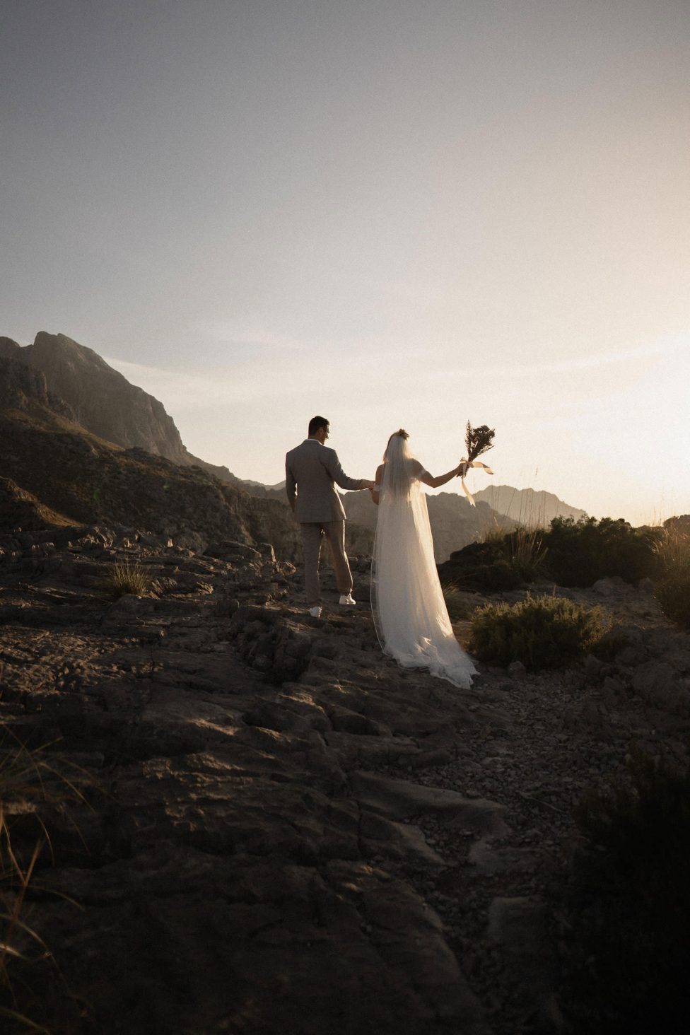 Hochzeitswahn - Ein Paar in Hochzeitskleidung hält Händchen auf einem felsigen Pfad inmitten von Bergen, während die Braut einen Blumenstrauß hält und die untergehende Sonne einen warmen Schein wirft.