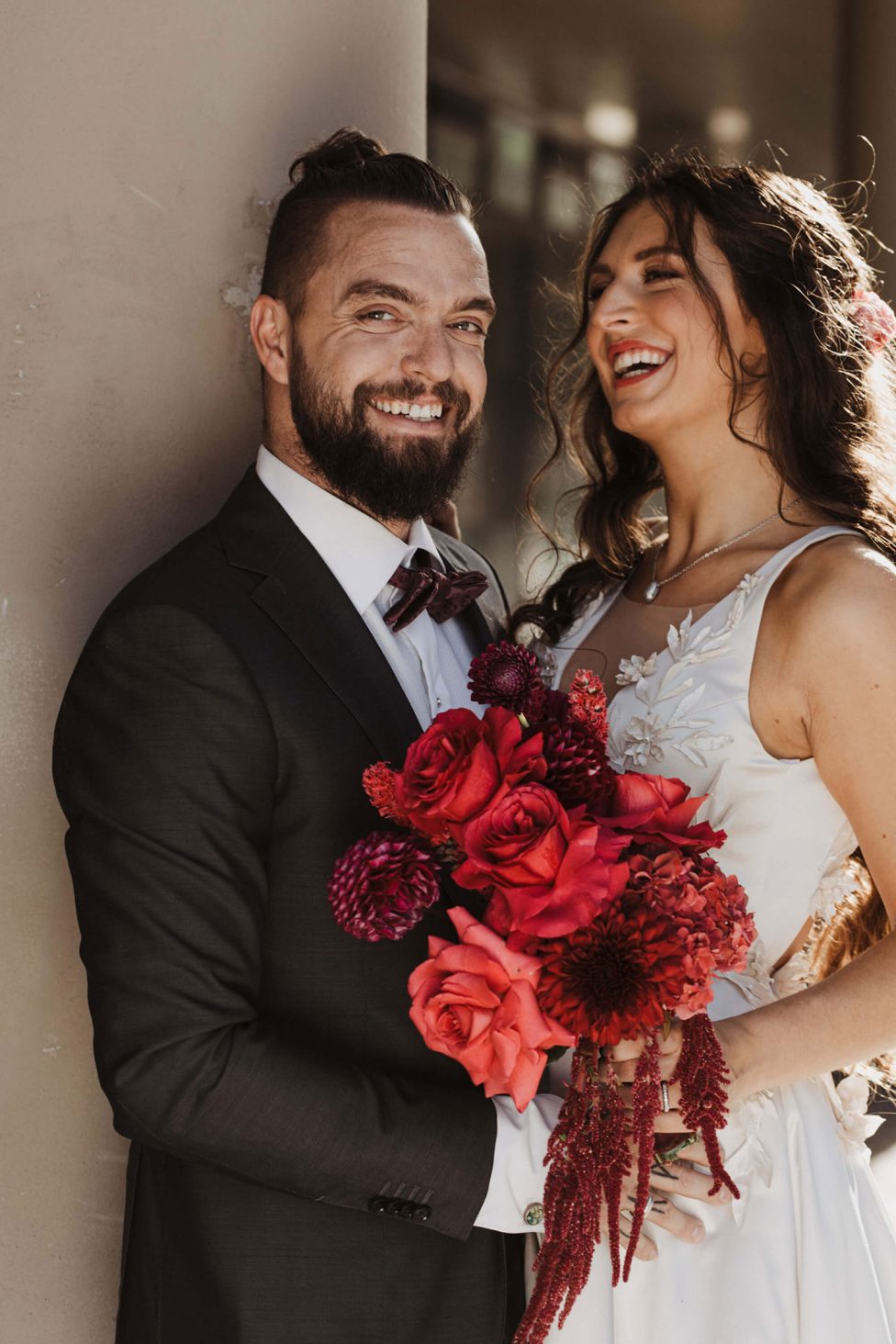 Hochzeitswahn - Ein strahlendes Paar an seinem Hochzeitstag, mit dem Bräutigam in einem klassischen Anzug, der die Braut in einem Spitzenkleid hält, die einen Strauß leuchtend roter Blumen in der Hand hält und einen freudigen Moment teilt.