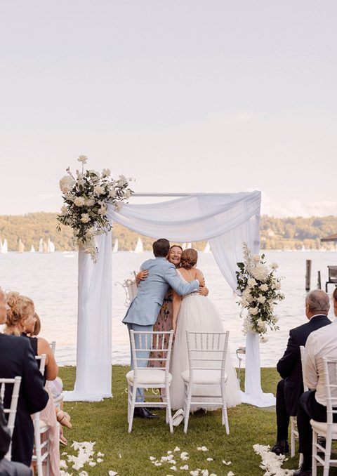 Hochzeitswahn - Eine Braut und ein Bräutigam umarmen sich vor einem See.