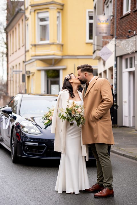 Hochzeitswahn - Ein Brautpaar küsst sich vor einem Porsche.