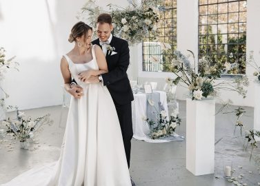 Hochzeitswahn - Eine Braut und ein Bräutigam umarmen sich in einem weißen Raum mit Blumen.
