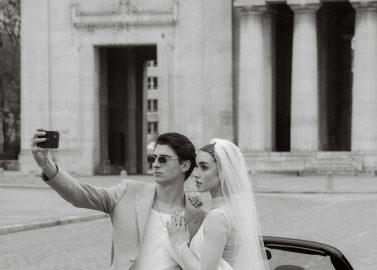 Hochzeitswahn - Ein stilvolles Paar macht ein Selfie mit der Braut in einem weißen Kleid und Schleier und dem Bräutigam in einem beigen Anzug neben einem eleganten Cabrio und fängt einen schicken, feierlichen Moment ein.