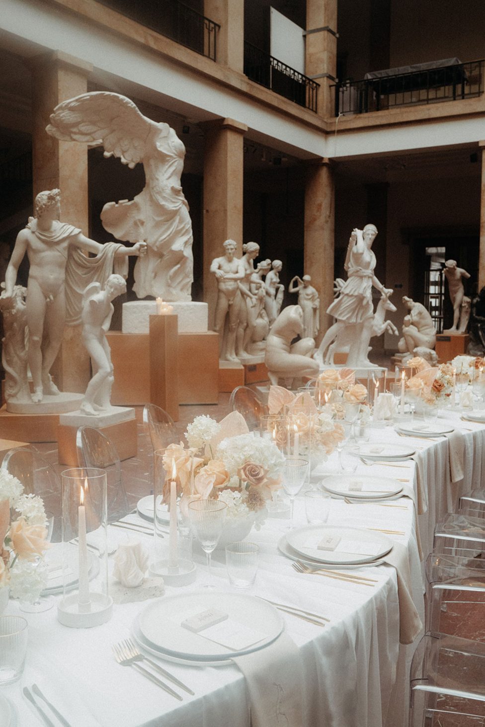 Hochzeitswahn - Ein elegantes Esszimmer mit transparenten Stühlen und Tischen, die mit Kerzen und Blumen geschmückt sind und zwischen klassischen Statuen in einem großen Museumssaal positioniert sind und ein Ambiente schaffen, das historische Kunst mit zeitgenössischem Veranstaltungsdesign verbindet.