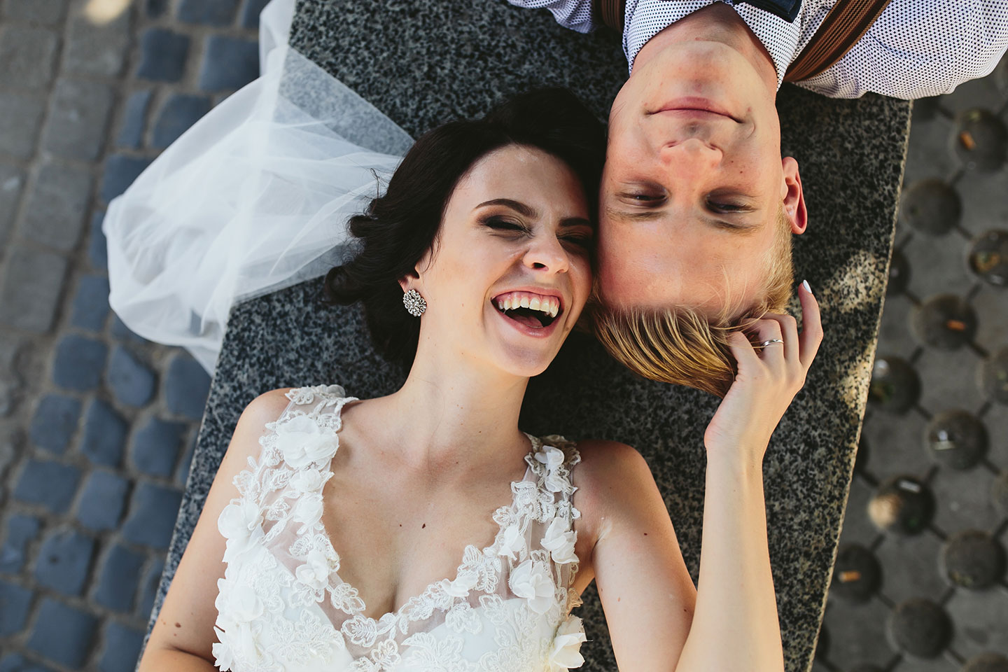 Hochzeitswahn - Braut und Bräutigam liegen auf einer Kopfsteinpflasterstraße, die Köpfe zusammengelegt, und lächeln freudig. Die Braut trägt ein weißes Spitzenbrautkleid, der Bräutigam ein Hemd mit Hosenträgern. Die Kulisse lässt einen verspielten, romantischen Moment erahnen.