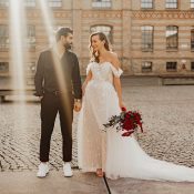 Hochzeitswahn - Ein frisch verheiratetes Paar, ein Mann im schwarzen Anzug und eine Frau im schulterfreien weißen Brautkleid, halten Händchen und lächeln sich in einem kopfsteingepflasterten Innenhof mit einem historischen Gebäude im Hintergrund an. Koenigliche-Porzellan-Manufaktur
