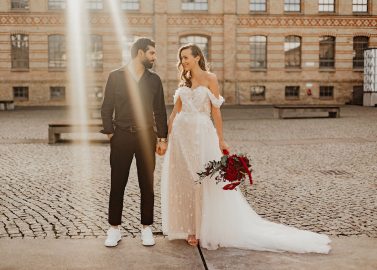 Hochzeitswahn - Ein frisch verheiratetes Paar, ein Mann im schwarzen Anzug und eine Frau im schulterfreien weißen Brautkleid, halten Händchen und lächeln sich in einem kopfsteingepflasterten Innenhof mit einem historischen Gebäude im Hintergrund an. Koenigliche-Porzellan-Manufaktur
