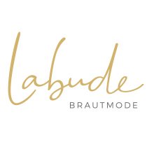 Hochzeitswahn - Das Bild zeigt das elegante Logo von „Labude Brautmode“, wobei „Labude“ in luxuriöser goldener Kursivschrift wiedergegeben ist. Darunter steht „BRAUTMODE“ in einer edlen grauen serifenlosen Schrift, alles vor einem makellos weißen Hintergrund. Landhotel-Voshoevel