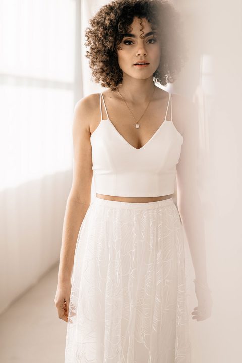Hochzeitswahn - Eine Person mit lockigem Haar steht drinnen und trägt ein ärmelloses weißes bauchfreies Top und einen hoch taillierten Rock mit einem zarten Spitzenmuster. Das natürliche Licht fällt durch die durchsichtigen weißen Vorhänge im Hintergrund und schafft eine mühelos schicke Atmosphäre, die direkt aus einem Auto Draft stammen könnte. Landhotel-Voshoevel