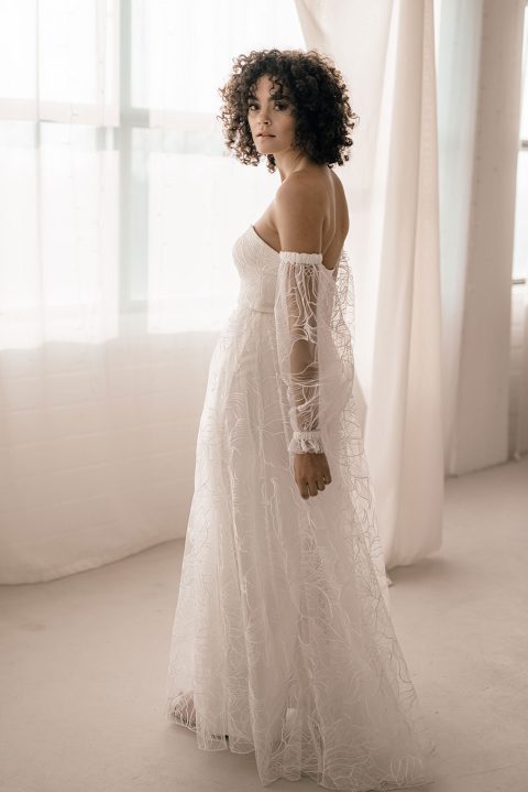 Hochzeitswahn - Eine Frau mit lockigem Haar, die ein schulterfreies weißes Brautrock-Kleid mit transparenten, langen Ärmeln trägt, steht in einem hellen Raum mit großen Fenstern, die mit hellen Vorhängen verhangen sind. Sie blickt über ihre Schulter in Richtung der Carson-Kamera.