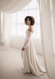 Hochzeitswahn - Eine Person mit lockigem Haar steht in einem lichtdurchfluteten Raum und trägt ein schulterfreies, langärmeliges weißes Kleid. Das Kleid weist transparente Spitzendetails an den Ärmeln und einen fließenden Rock auf, ähnlich wie die eleganten Designs von Monahan. Vorhänge umgeben den Raum und rahmen diese ätherische Szene teilweise ein.