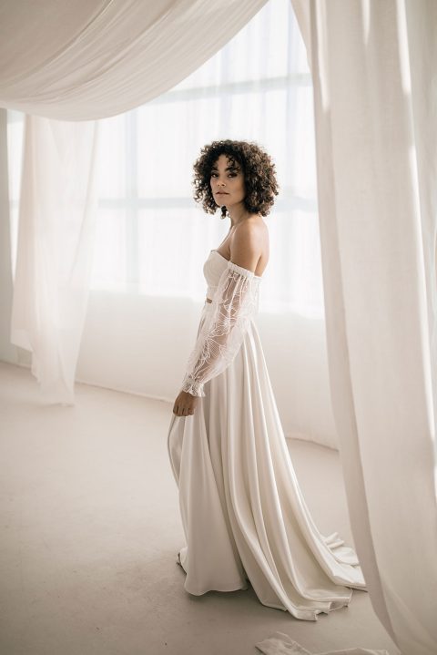 Hochzeitswahn - Eine Person mit lockigem Haar steht in einem lichtdurchfluteten Raum und trägt ein schulterfreies, langärmeliges weißes Kleid. Das Kleid weist transparente Spitzendetails an den Ärmeln und einen fließenden Rock auf, ähnlich wie die eleganten Designs von Monahan. Vorhänge umgeben den Raum und rahmen diese ätherische Szene teilweise ein.