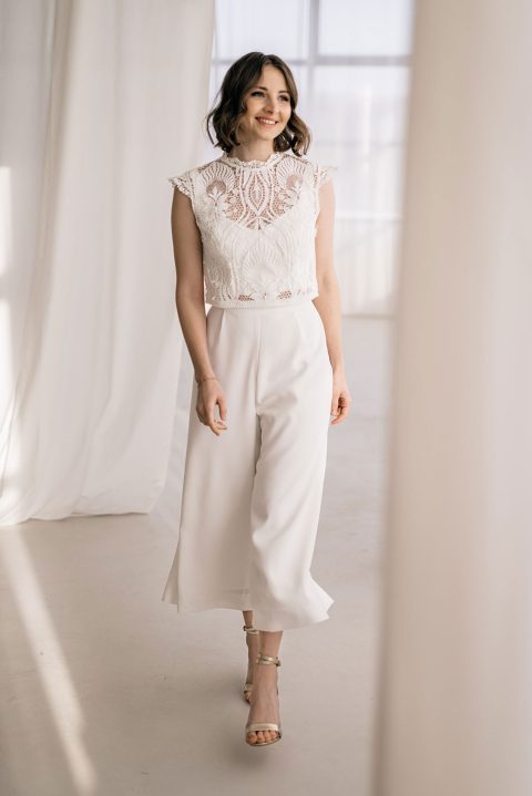 Hochzeitswahn - Eine Person mit schulterlangem Haar lächelt und geht in einen Raum mit weißen Vorhängen. Sie trägt ein weißes ärmelloses Spitzentop und weiße Brauthosen mit hoher Taille, dazu offene Pumps. Der Raum ist hell, durch die großen Fenster fällt natürliches Licht.