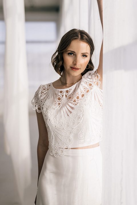 Hochzeitswahn - Eine Frau, die an Juno erinnert, steht drinnen mit erhobenem rechten Arm, der an einen weißen Vorhang gelehnt ist. Sie trägt ein weißes Outfit mit aufwendigen Spitzendetails am Oberteil, ähnlich einem zarten Brauttopper. Der sanft beleuchtete Hintergrund schafft eine saubere und luftige Atmosphäre. Ihr kurzes, welliges braunes Haar umrahmt ihr Gesicht, während sie in die Kamera blickt.
