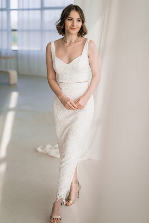 Hochzeitswahn - Selma steht neben einem weißen Vorhang und trägt ein ärmelloses Brautrock-Spitzenkleid mit tiefem Ausschnitt und Saum bis zur Mitte der Wade. Dazu trägt sie beige Sandalen mit hohen Absätzen. Im Hintergrund sind große Fenster mit transparenten Vorhängen zu sehen, die den Raum mit natürlichem Licht füllen.