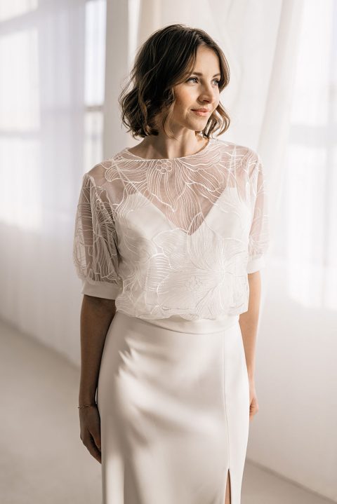 Hochzeitswahn - Eine Person mit schulterlangem braunem Haar trägt einen weißen, halbtransparenten Brauttopper über einem taillierten weißen Rock mit kleinem Schlitz. Sie steht in einem sanft beleuchteten Raum mit transparenten weißen Vorhängen und blickt mit einem leichten Lächeln zur Seite.