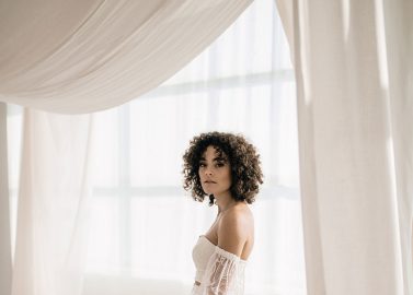 Hochzeitswahn - Eine Frau mit lockigem Haar trägt ein weißes, schulterfreies Brautkleid mit Spitzenärmeln. Sie steht in einem sanft beleuchteten Raum, um den sie hing, und die durchsichtigen weißen Vorhänge verleihen ihr fast automatisch einen Hauch von Eleganz. Der helle Boden passt zur insgesamt minimalistischen und raffinierten Ästhetik. Landhotel-Voshoevel