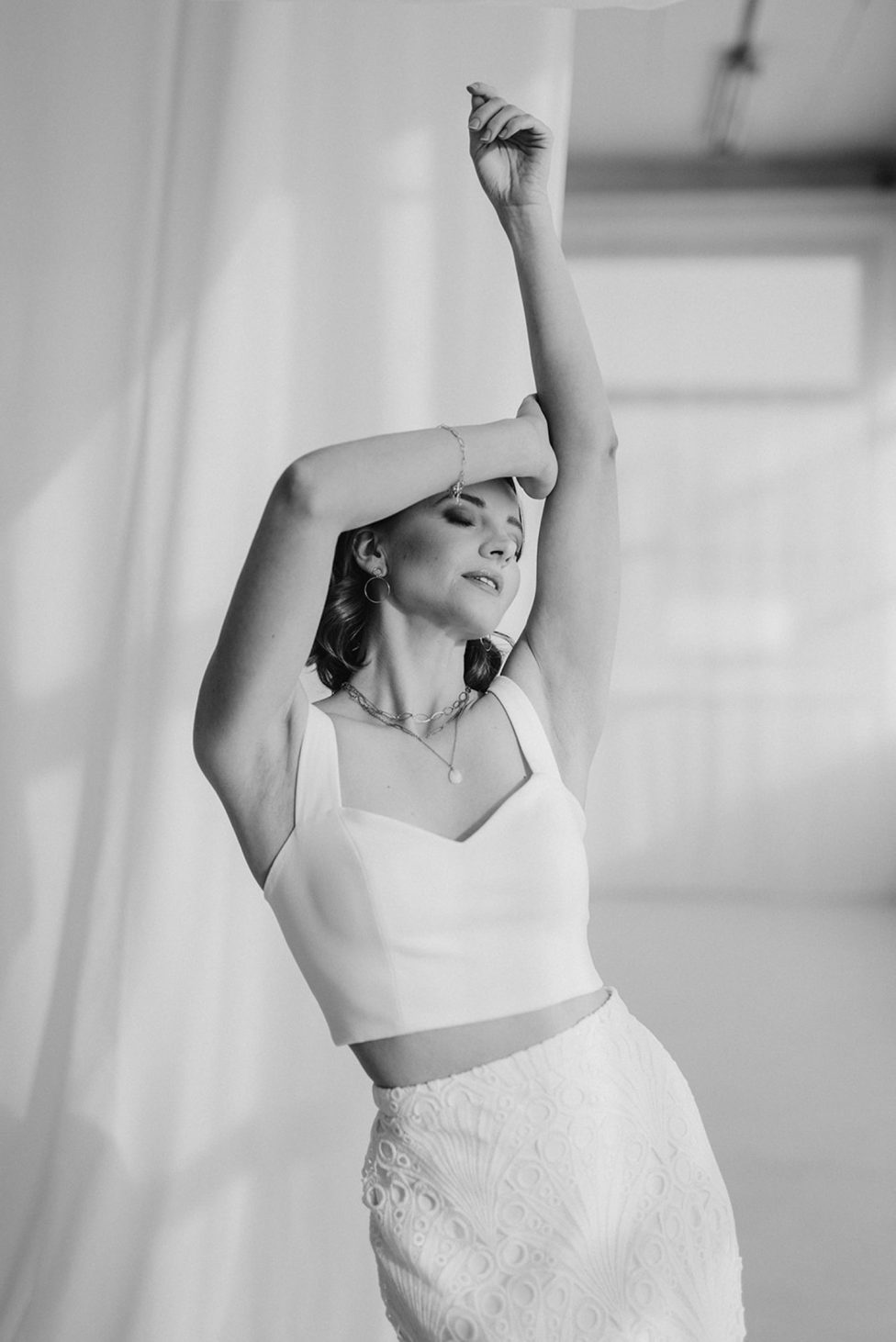 Hochzeitswahn - Schwarzweißfoto einer Frau in einem weißen ärmellosen Oberteil und einem strukturierten Rock. Sie posiert mit einem Arm über dem Kopf gebeugt und dem anderen erhoben, wobei sie leicht nach oben blickt. Der Hintergrund, der einer automatischen Skizze ähnelt, zeigt eine weiche, verschwommene Kulisse mit drapiertem Stoff. Landhotel-Voshoevel