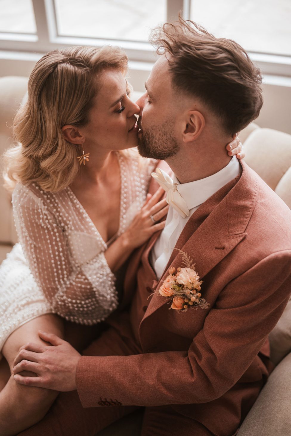 Hochzeitswahn - Ein Paar sitzt eng beieinander und küsst sich. Die Frau in einem weißen Kleid mit transparenten, gepunkteten Ärmeln trifft den Mann in einem rosafarbenen Anzug, der mit einer Ansteckblume geschmückt ist. In einer intimen Innenumgebung am Fenster ist es, als könnte ihre Liebesgeschichte ihren eigenen Entwurf schreiben. Hochzeit in der Kulturfabrik Coburg.