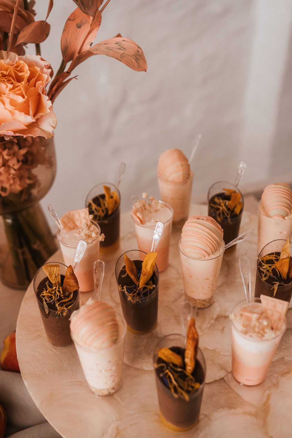 Hochzeitswahn - Auf einem runden Marmortablett stehen verschiedene kleine Dessertbecher, einige mit Schokoladenmousse und andere mit einer hellen Cremefüllung. Jeder Becher ist mit Garnituren wie Trockenfrüchten dekoriert und wird von einem kleinen Löffel begleitet. Im Hintergrund steht eine Vase mit rosa Blumen, die der Szene einen Hauch von Eleganz verleiht. Hochzeit in der Kulturfabrik Coburg.