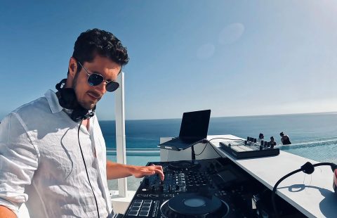 Hochzeitswahn - DJ Fabian Baroud, in weißem Hemd und Sonnenbrille, legt draußen am Meer Musik auf. Mit seinem Mixer und Laptop-Setup zieht er die Menge in seinen Bann. Der klare Himmel und das Meer bilden eine atemberaubende Kulisse, während einige Leute in der Ferne die Musik genießen. Hochzeit in der Kulturfabrik Coburg.