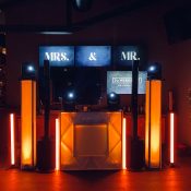 Hochzeitswahn - Bei einer Veranstaltung ist ein DJ-Pult mit orange-weißer Beleuchtung aufgebaut. Hinter dem Pult zeigen Bildschirme die Worte „MRS. & MR.“. Auf dem rechten Bildschirm steht „Die WERKS-Probe“. DJ Fabian Baroud rockt den Veranstaltungsort, während das Pult von großen Lautsprechern und beleuchteten Säulen umgeben ist. Hochzeit in der Kulturfabrik Coburg.