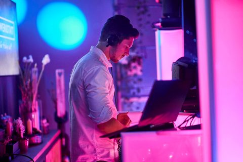 Hochzeitswahn - Eine Person mit Kopfhörern steht hinter einem Laptop in einem von rosa und blauem Licht erhellten Raum und sieht aus, als würde sie Fabian Barouds DJ-Vibes kanalisieren. Die Person scheint auf den Laptop-Bildschirm fokussiert zu sein, während im Hintergrund verschiedene Objekte zu sehen sind, darunter ein Fernseher, Pflanzen und Dekorationen. Hochzeit in der Kulturfabrik Coburg.